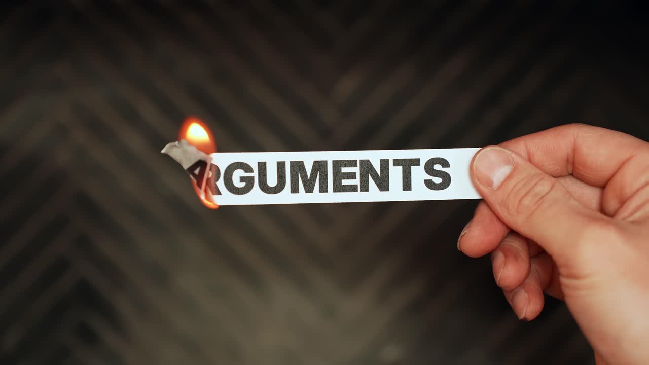 Hand burns a paper strip reading “ARGUMENTS” with flame, symbolizing conflict, anger, relationship problems, and heated disputes. Dark background concept b-roll