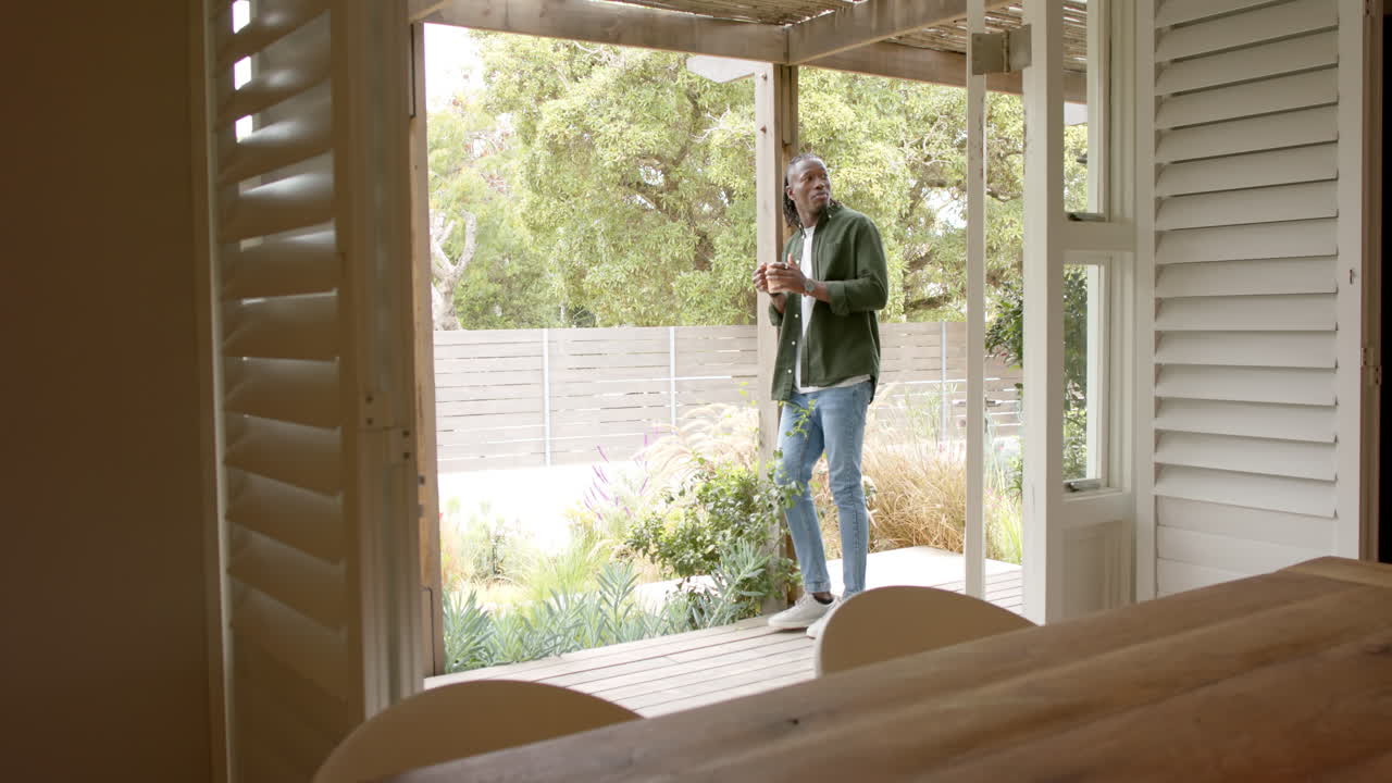 Standing on porch, African american man enjoying coffee and looking at garden scenery