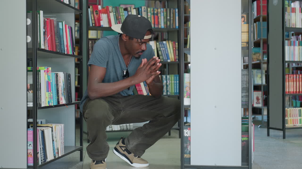Um jovem estudante vestindo uma camiseta casual com óculos de sol na camisa, agachado no corredor da biblioteca, inclina-se em direção à prateleira inferior enquanto examina os títulos dos livros, busca focada sob luz interna suave e paredes verde-azuladas