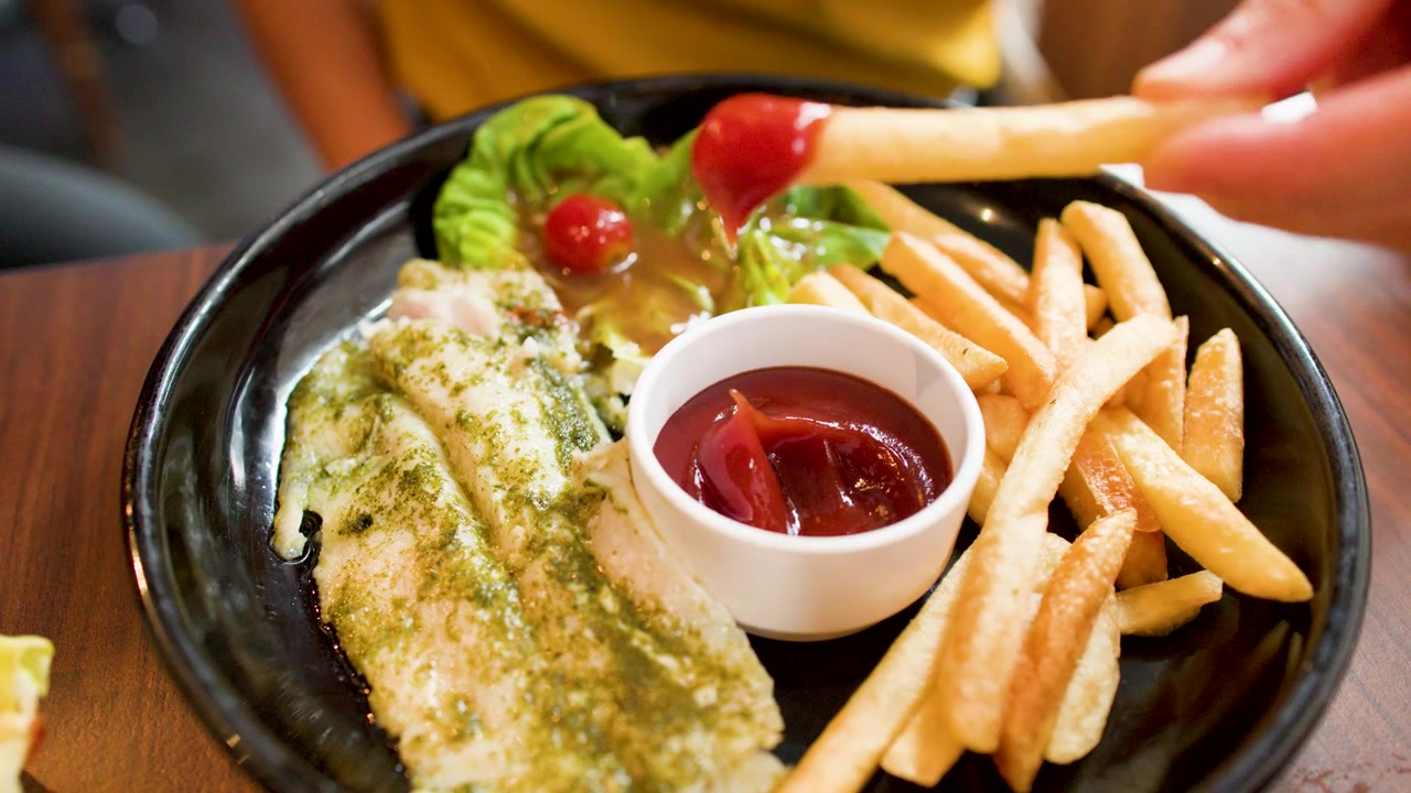 Una mano intinge una patatina fritta dorata nel ketchup rosso accanto a pesce alla griglia con pesto.