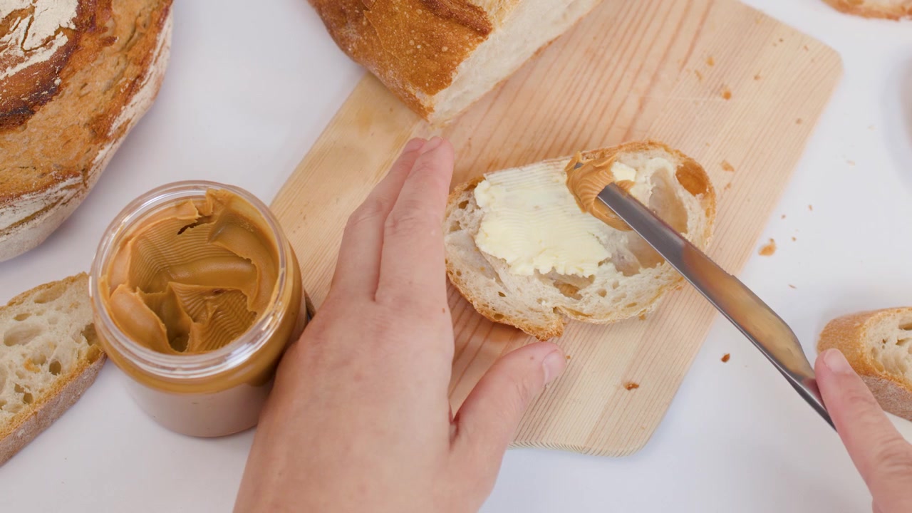 Close-up overhead shot of hands spreading peanut butter onto toasted artisan bread on wooden board.