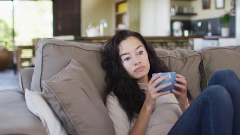 relaxed biracial woman with vitiligo drinking coffee, sitting on sofa