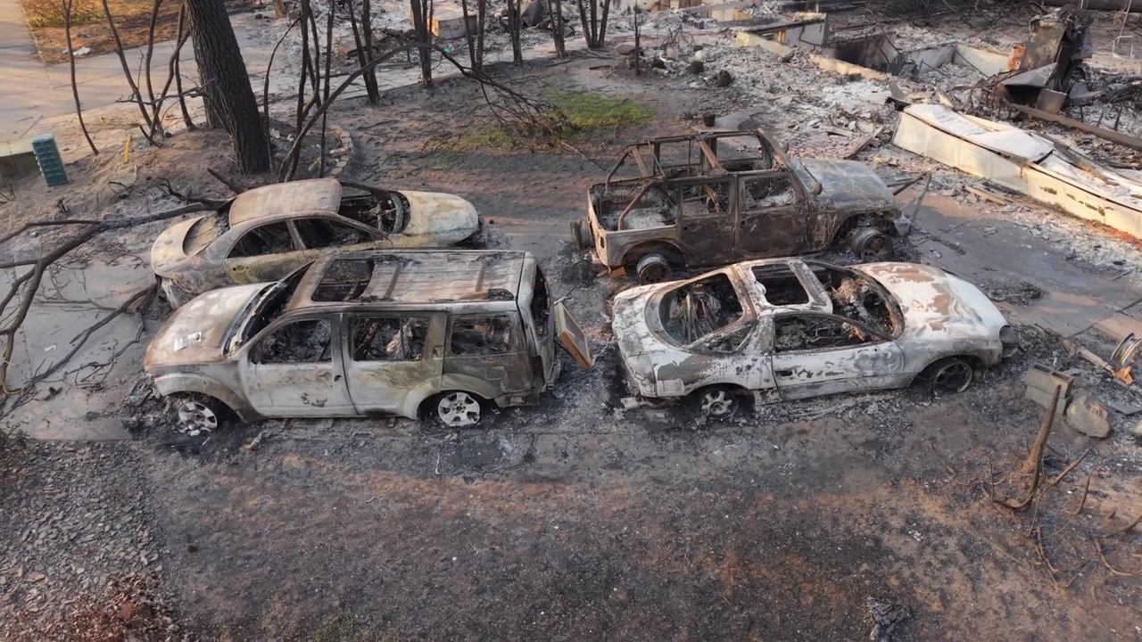 Danni dell'incendio Spokane Old Trails mostrati in un'orbita aerea sopra quattro auto bruciate su un lotto residenziale distrutto, circondato da detriti e distruzione causata da incendi boschivi