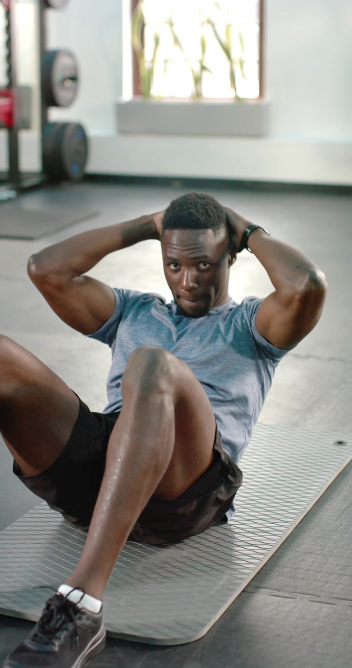 Vertical video: Placing hands behind head, man performing crunches on mat in gym for core strength