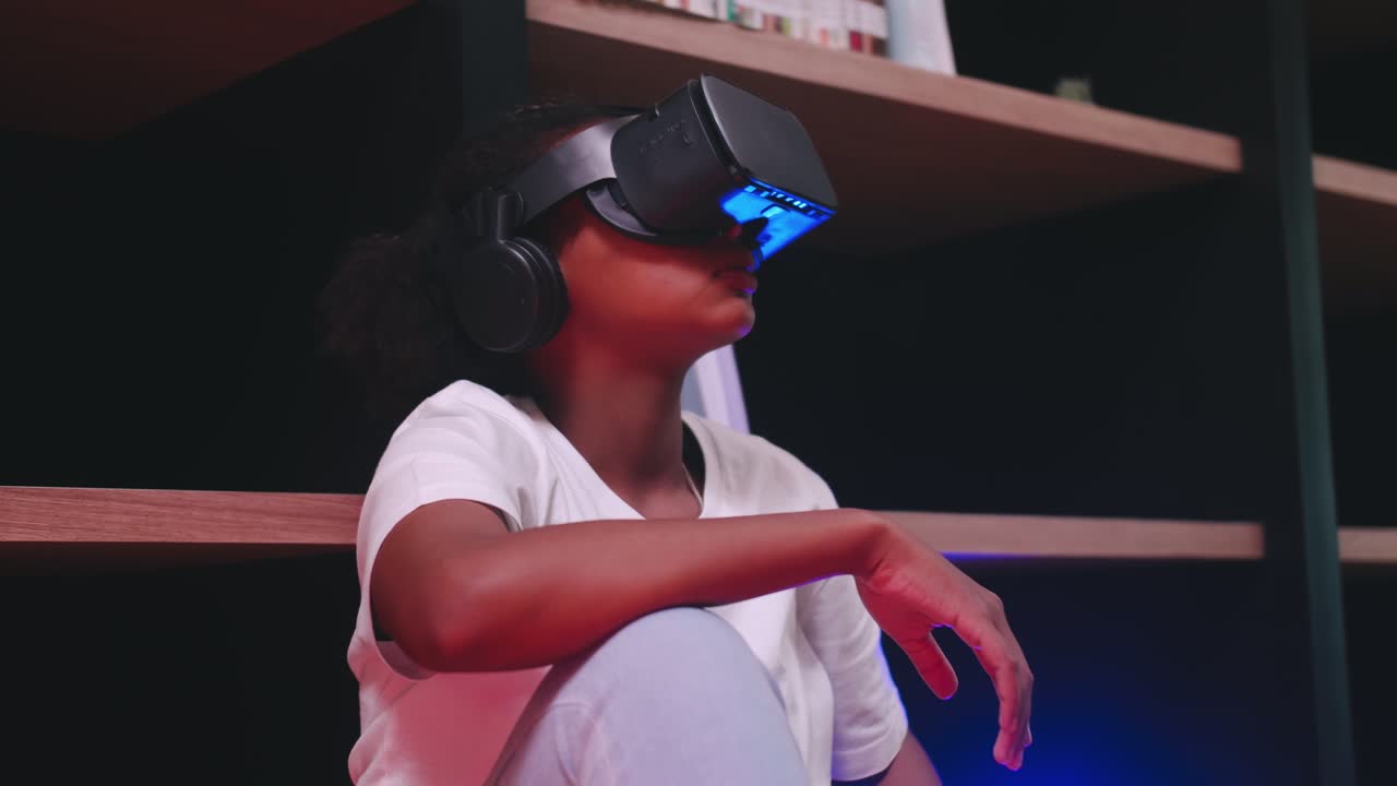 A schoolgirl sitting in front of library's bookshelf and using virtual reality glasses.