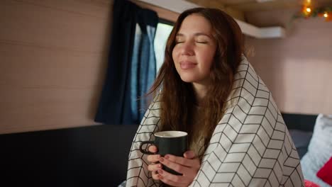 Close-up-shot-of-a-happy-brunette-girl-holding-a-black-mug-of-tea-in-her-hands-and-enjoying-the-aroma-and-warmth-of-the-drink,-wrapped-in-a-large-white-blanket-Sitting-in-a-trailer-in-a-camp-during-her-picnic-outside-the-city-in-the-summer