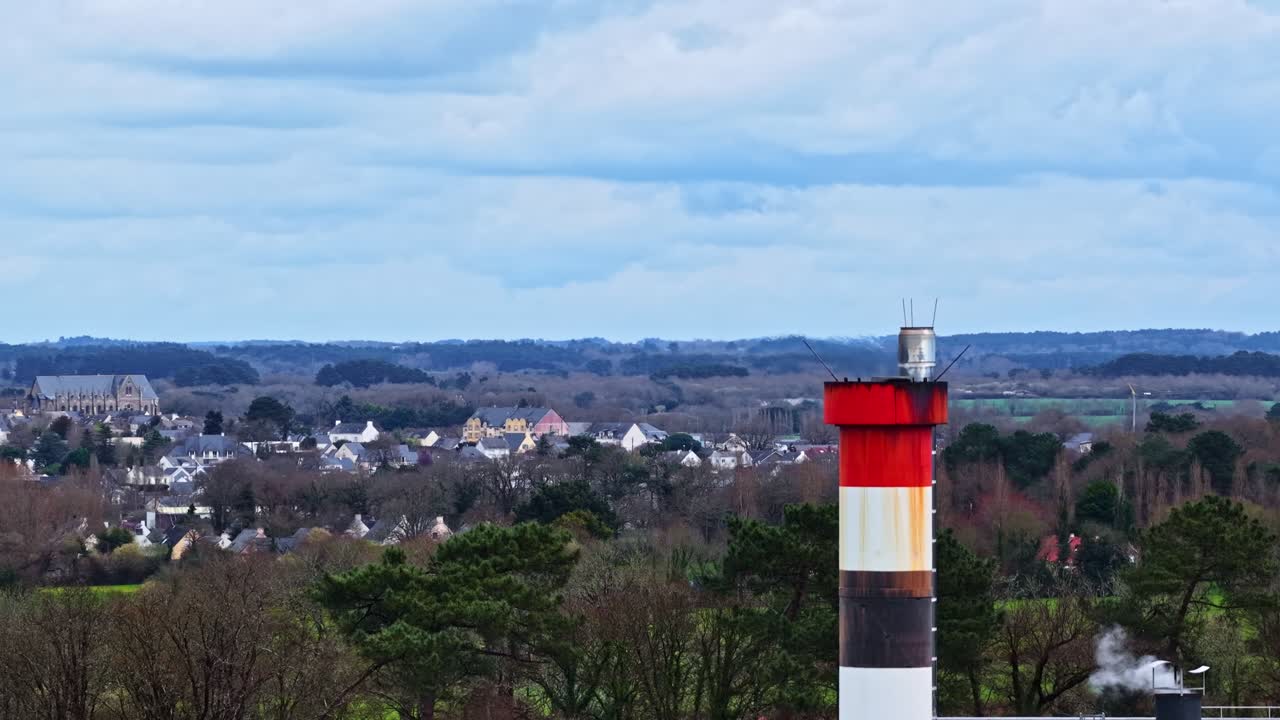 Luftaufnahme eines kleinen Dorfes in grüner ländlicher Umgebung, mit einem Fabrikschornstein im Vordergrund.