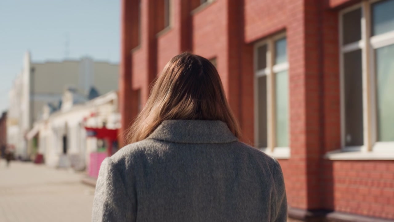 Pretty caucasian woman walking past brick building, commuter mood, gray coat, back view, soft sunlight, winter city street, reflective pace, casual fashion details, distant storefronts