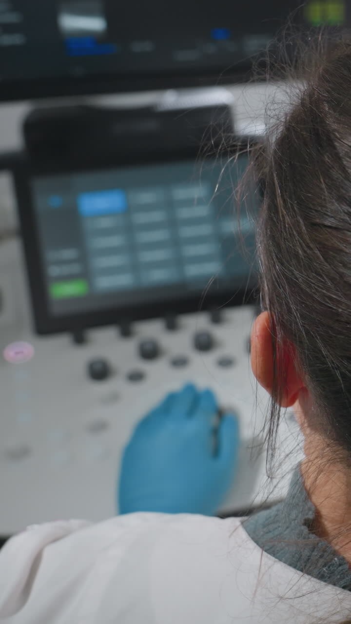 Kliniker bagfra ved ultralydsskærm og konsol tæt på handskebeklædt hånd på betjeningsknapper med synlige skærmforudindstillinger, professionel ved arbejdsstation, der gennemgår billeddannelse og indstillinger, roligt klinikrum