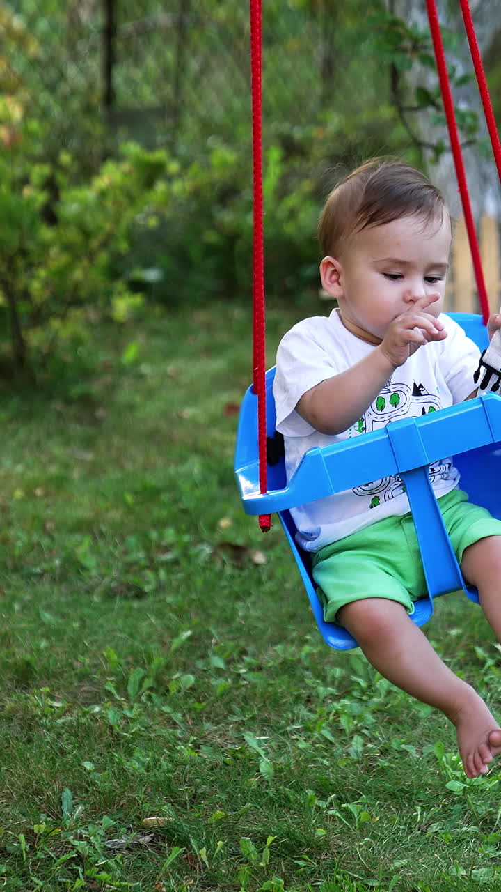 Peaceful Caucasian baby boy swaying in the blue swing in the garden. Trees and wooden fence at backdrop. Vertical video