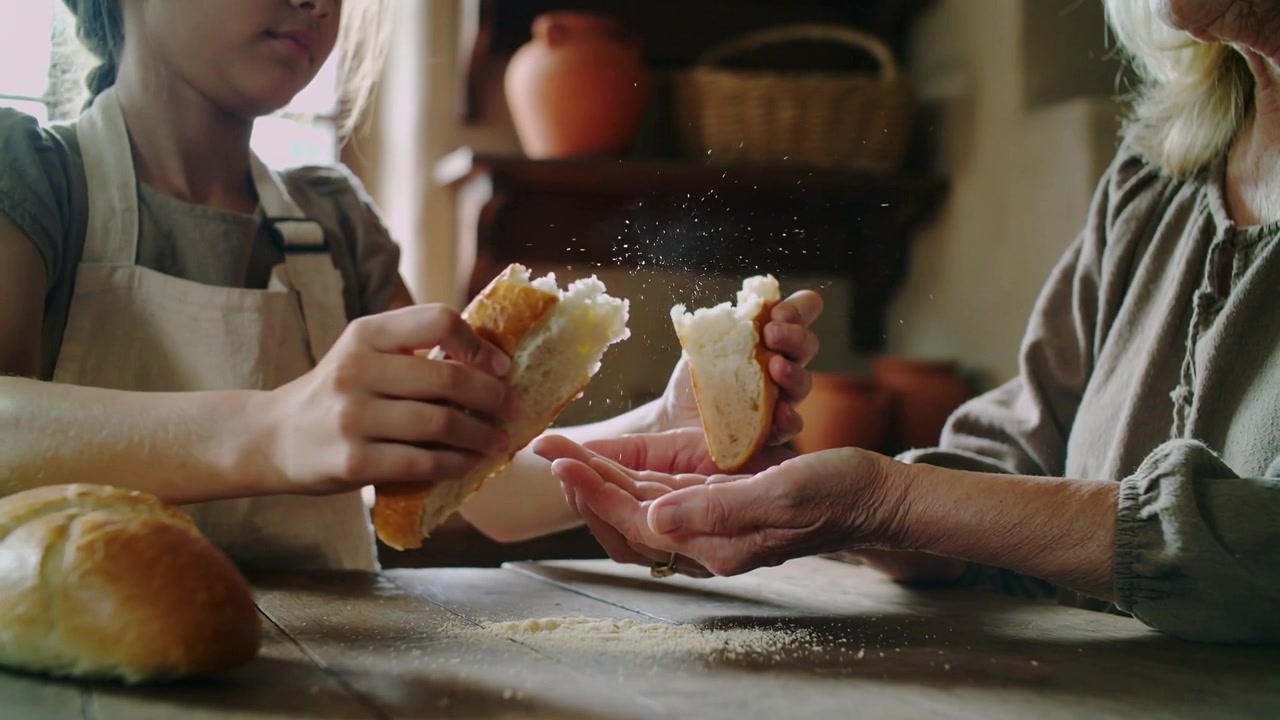 Pulling child wearing apron tearing loaf apart at rustic table, prompted by grandma's cupped hands