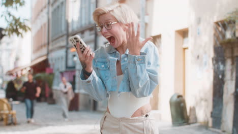 feliz mujer rubia caucásica usando teléfono inteligente celebrando ganar buenas noticias mensaje en la ciudad calle de la ciudad
