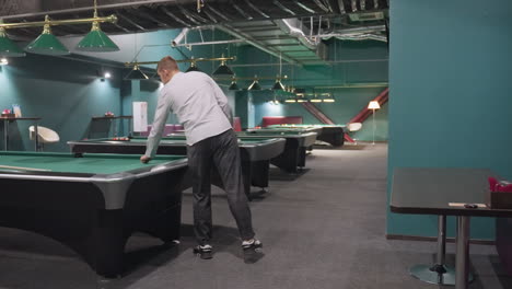 ack toma de un jugador con camisa blanca y pantalones grises entrando en la sala de billar, de pie frente a la mesa de billar verde, recogiendo la pelota blanca.