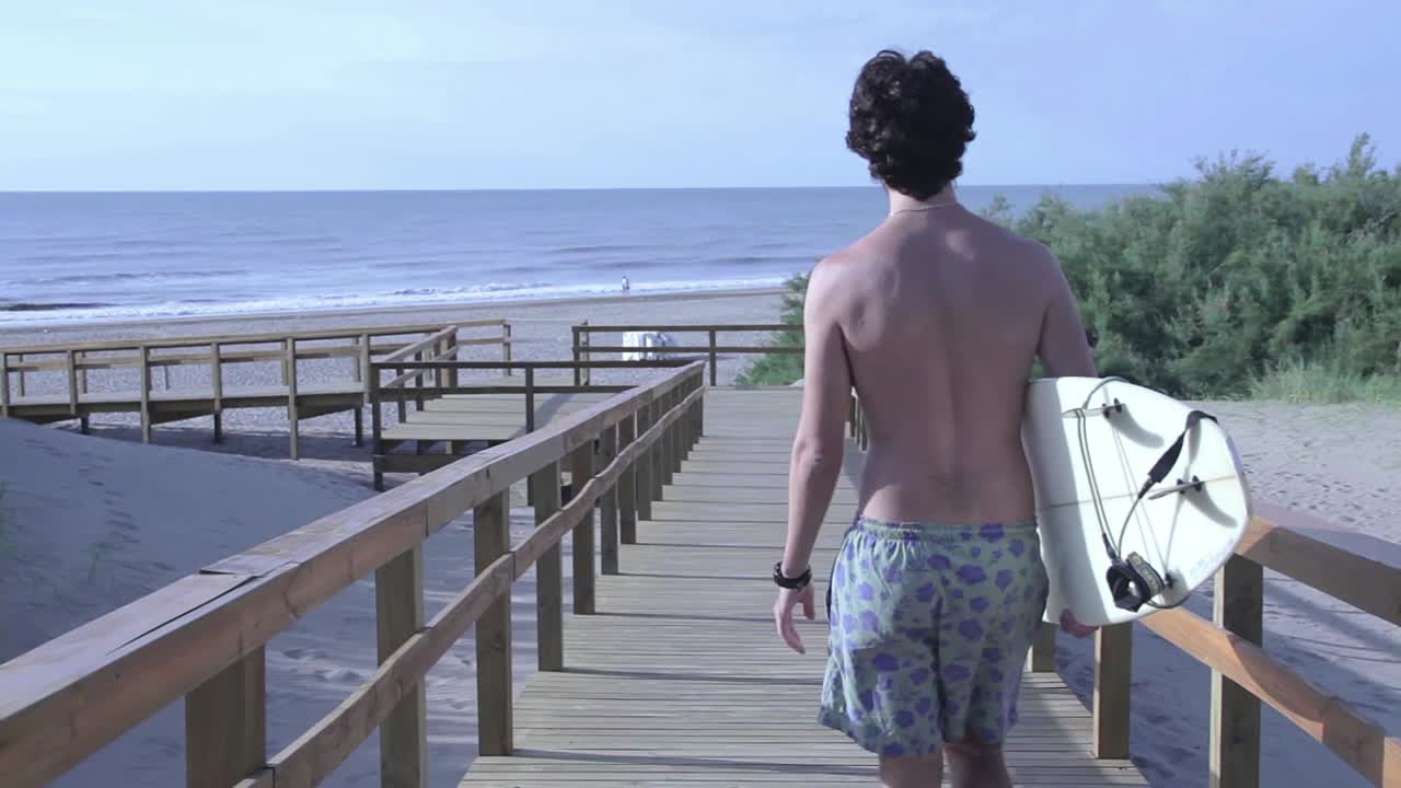 Young Man Walking on Boardwalk with Surfboard Towards the Ocean