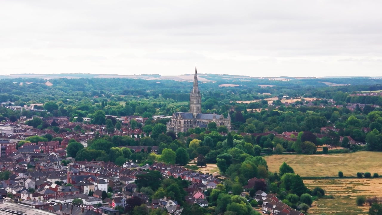 Riprese aeree con drone della Cattedrale di Salisbury e della sua alta guglia, circondata da alberi, strade storiche e la dolce campagna del Wiltshire.