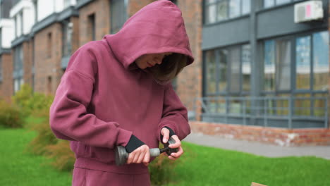 young female athlete in wine hoodie adjusting metal dumbbell outdoors beside brick building, ensuring secure grip before street workout amid courtyard greenery, symbolizing wellness perseverance