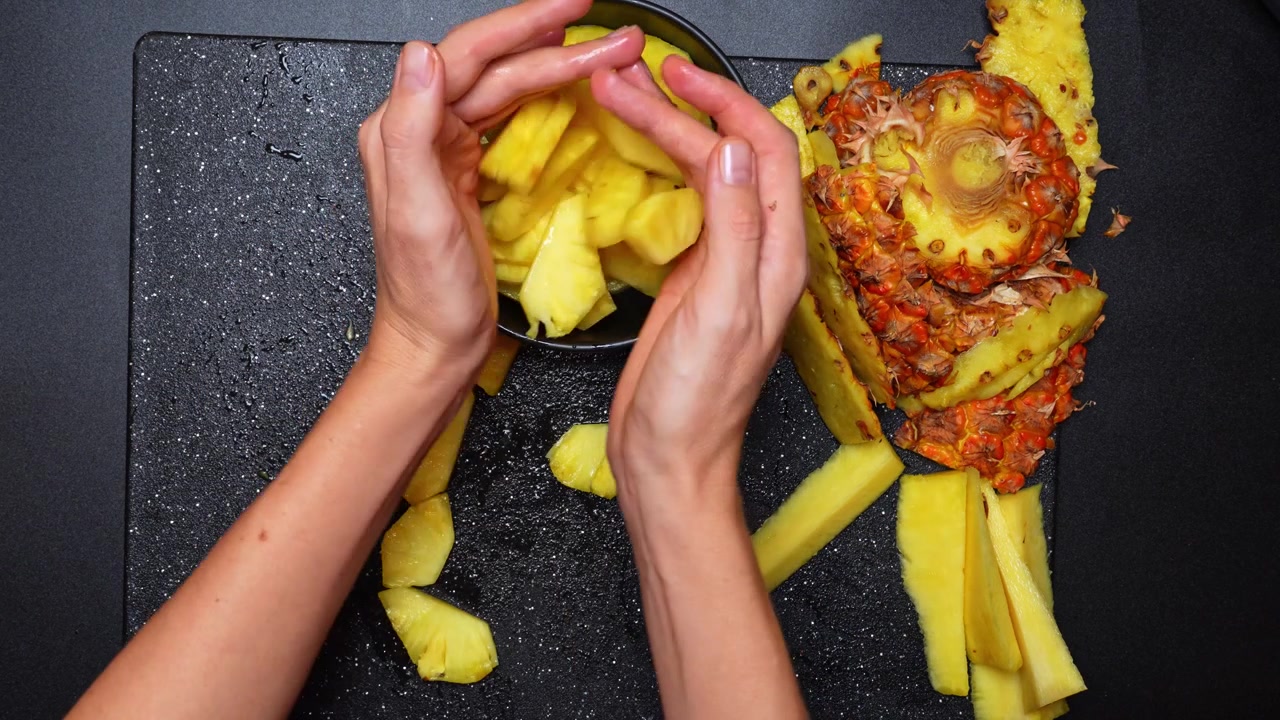 Cortando abacaxi fresco em uma tábua de corte preta com uma tigela por perto. Mãos cortando abacaxi fresco em pedaços em uma tábua de corte preta ao lado de uma tigela para a fruta