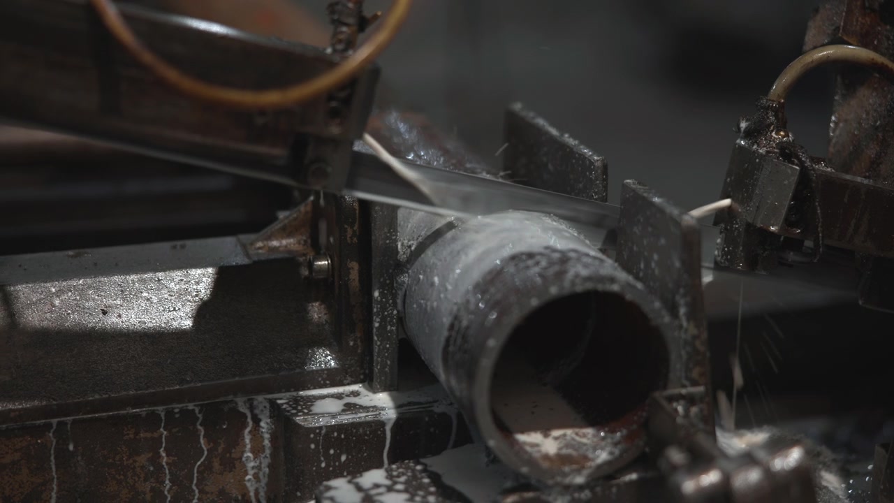 Industrial motion of horizontal bandsaw slicing clamped metal pipe in workshop, with coolant streaming across blade, guide plates, clamp, and steel surface during precise cutting process