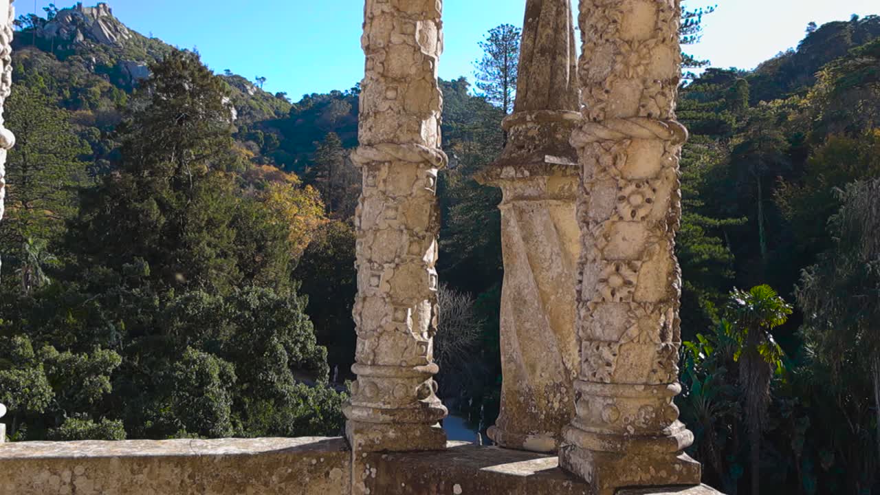 Vista panoramica dalla torre collinare del Palazzo Quinta da Regaleira con intagli complessi e colonne di pietra ornate che incorniciano giardini esotici e le colline di Sintra nella calda luce del sole. Esplorare un paesaggio mistico