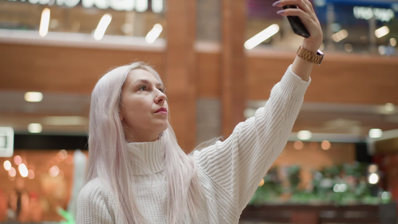 Leisurely female shopper sits on tiled bench beside indoor mall fountain raising mobile phone to pose capture fountain jets shimmer lights wearing sweater while shoppers stroll past displays