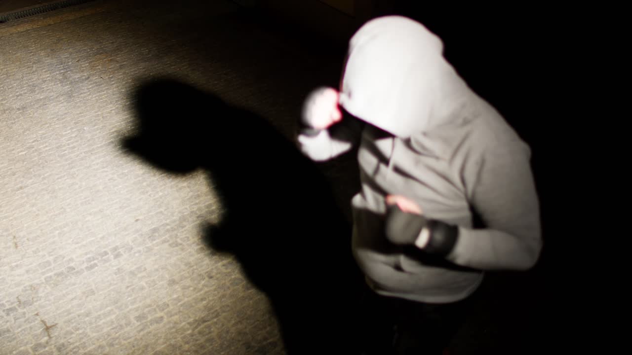 Hooded man shadow boxing under streetlight at night on a brick pavement.