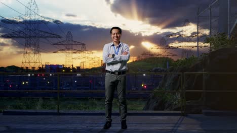 tomas de cuerpo entero de un trabajador profesional asiático de pie cerca de la torre de alto voltaje, instalación industrial, su amplia sonrisa a la cámara y cruzar el brazo sobre su pecho