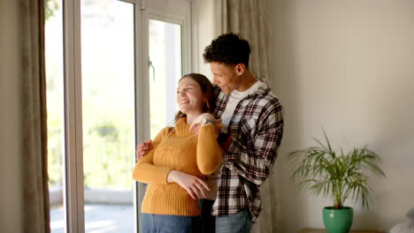 Happy-diverse-couple-looking-through-window-and-embracing-at-home,-in-slow-motion