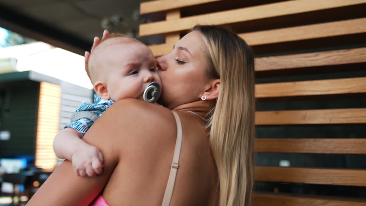 Young mom is waving her infant baby. Tiny boy suckling a pacifier lying on mother's shoulder.