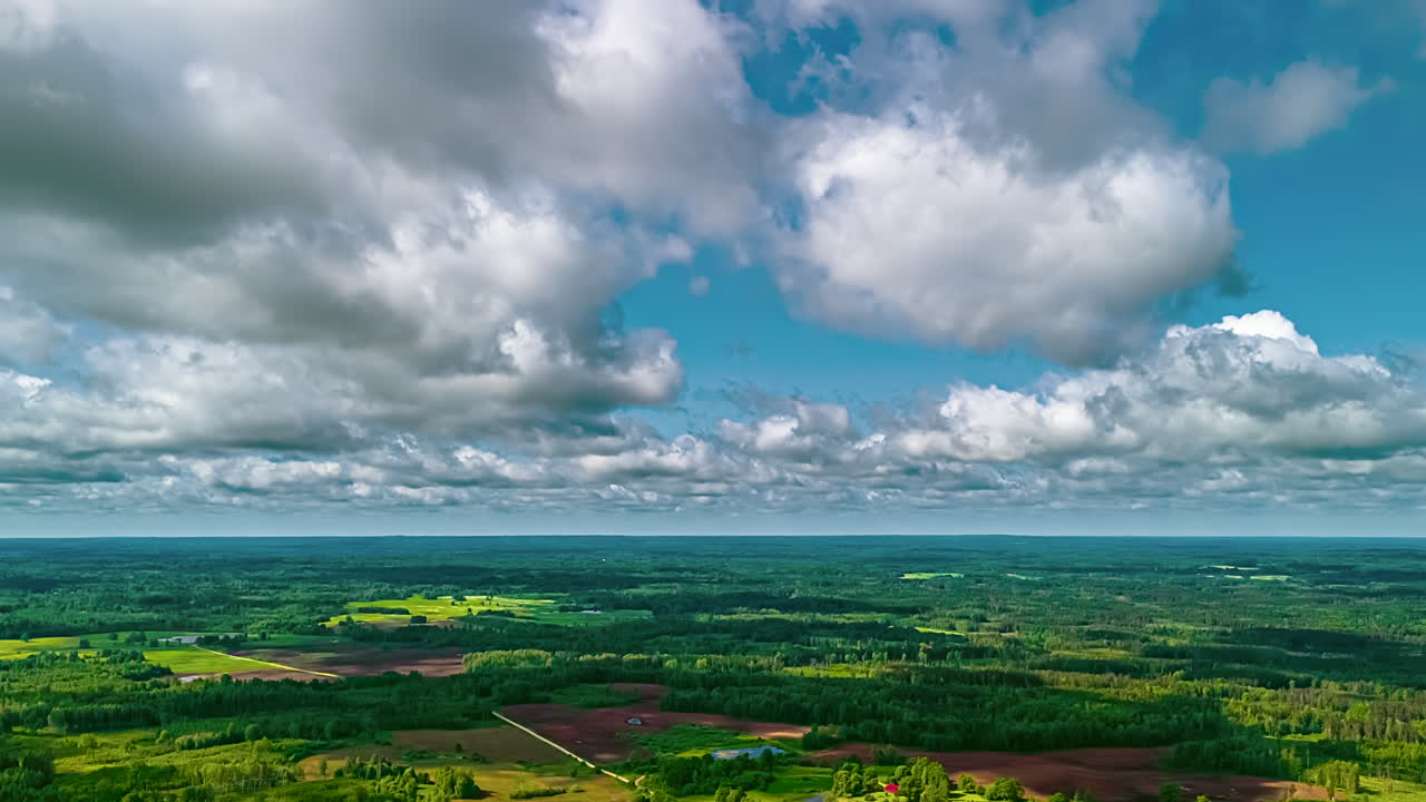 A majestic aerial time-lapse shows white clouds moving across the sky, casting beautiful sun rays, or god rays, over a vast and lush green forest landscape in Latvia