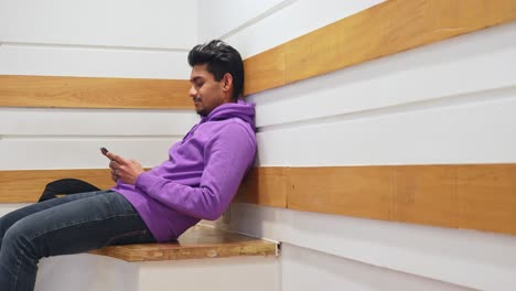 Guy-reading-on-his-phone-while-sitting-in-the-corner-of-the-room-and-leaning-his-back-against-the-wall,-static