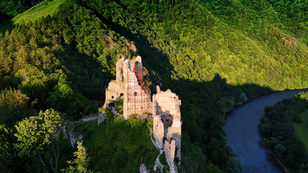 Approaching the ruins of the ancient castle lit by the bright sun. Amazing wooded Tatra mountains and narrow river at backdrop. Aerial view