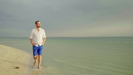 a young well-looking man in light clothes and glasses goes along the beach along the sea escape from