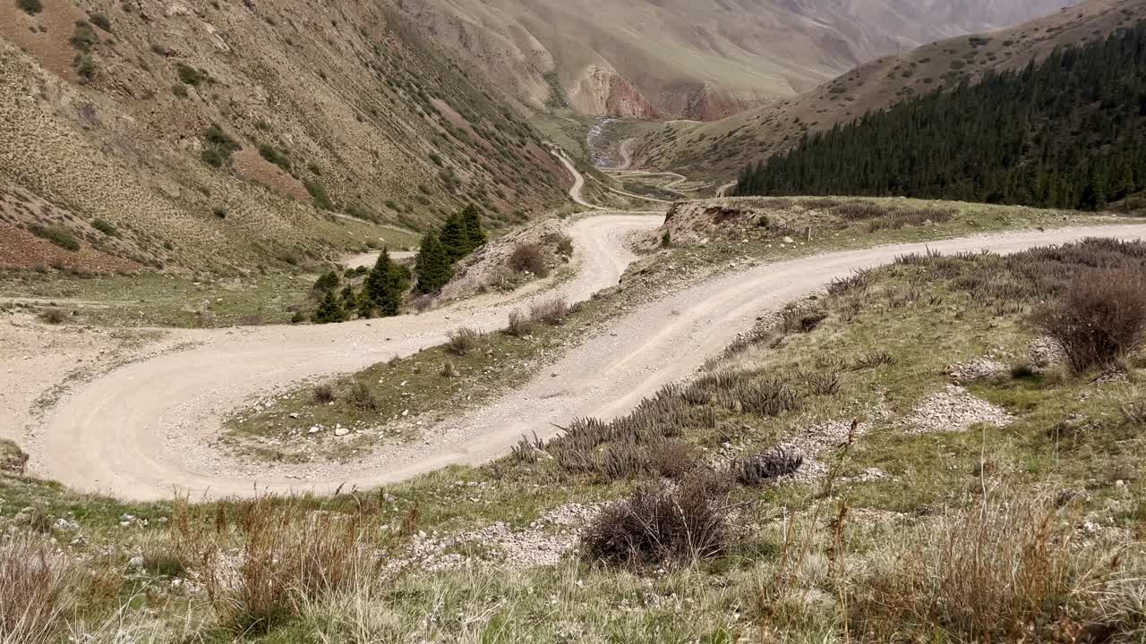 A motorcycle traveler navigates the famous winding switchbacks leading to Song-Kul Lake in Kyrgyzstan. The gravel road snakes through a vast mountain valley under a dramatic alpine landscape