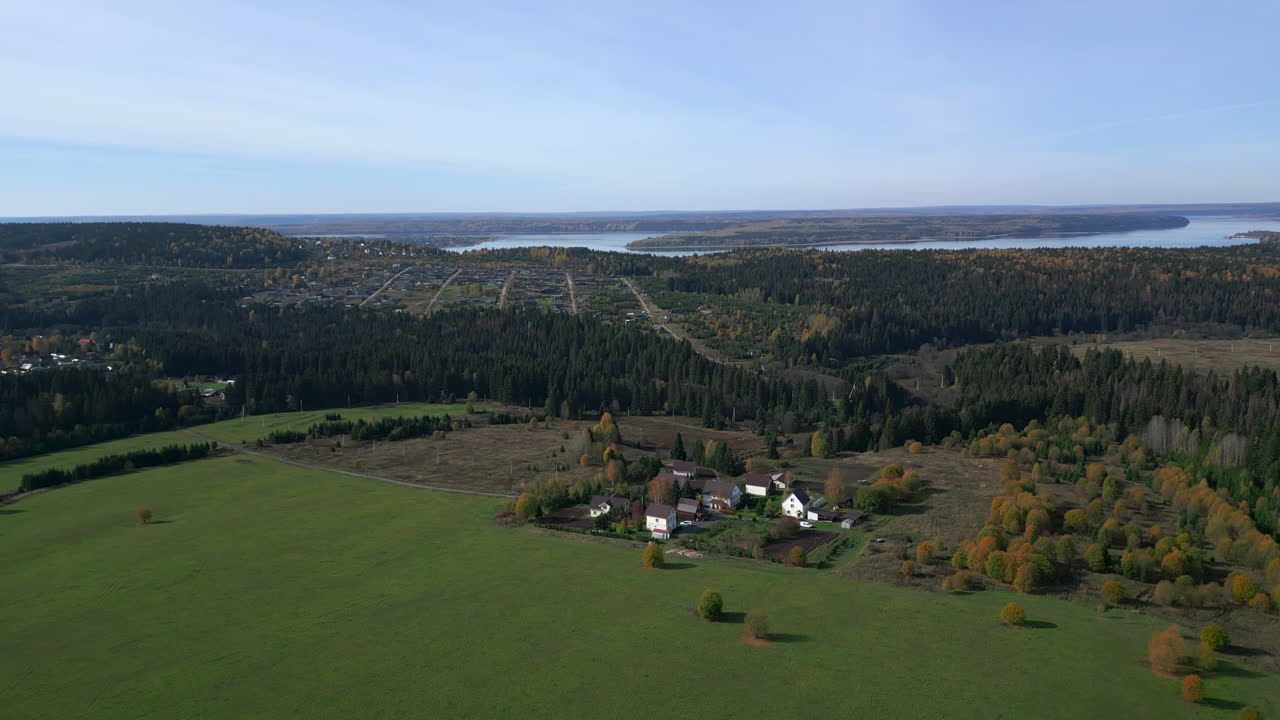 Ilmakuva vihreästä niittykylästä järven lähellä, kumpuilevaa metsää ja hajallaan olevia taloja syyspuiden kera, etäällä. Leike.