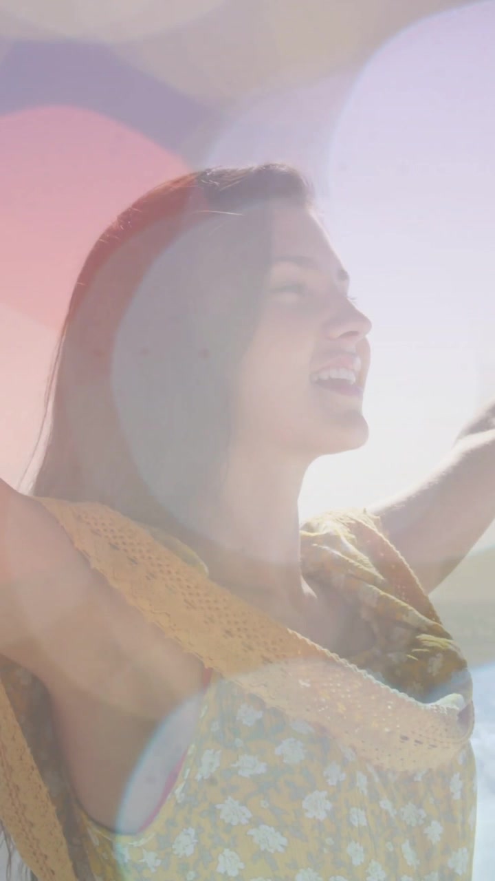 Dikey video: Güneş sarı elbiseli kadına vuruyor, kollarını kaldırarak sahilde güneşleniyor, deniz bokeh'i, mayo askısı.