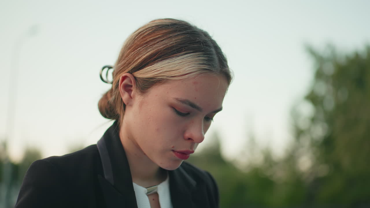Young lady with neatly parked hair to back appears deeply focused with soft expression while looking downward outdoors with blur background of green trees under clear bright sky