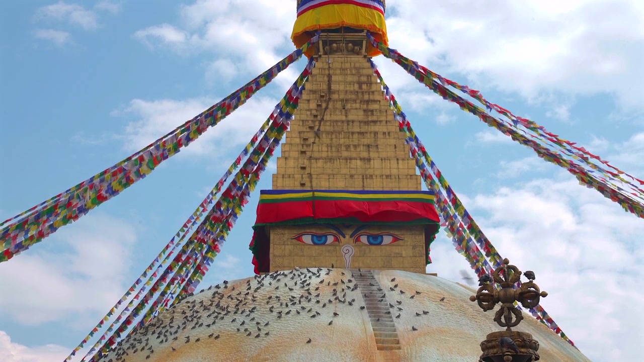 Un'inquadratura statica dal basso dell'iconico Stupa di Boudhanath, con gli occhi del Buddha, bandiere di preghiera colorate e piccioni sulla cupola bianca, contro un cielo blu a Kathmandu, Nepal.