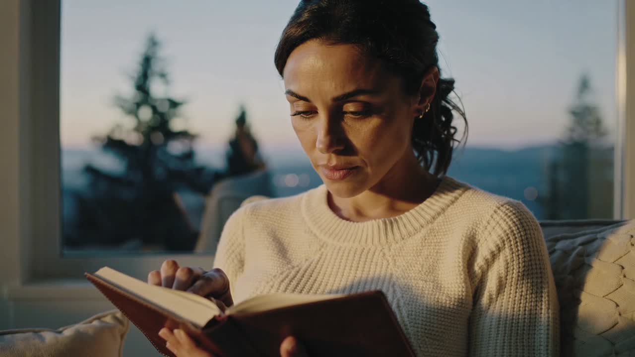 Donna che legge un libro con copertina rigida su un divano vicino a una grande finestra mentre la luce del giorno svanisce, indossando un maglione di lana