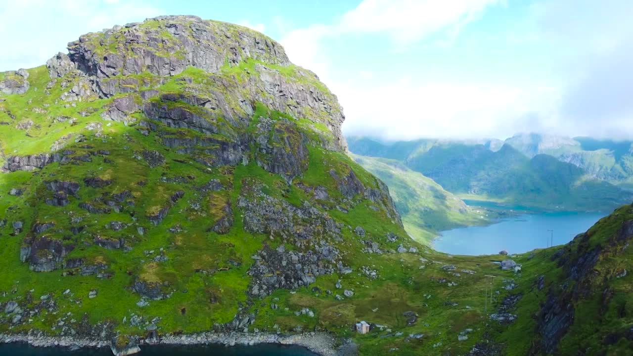 Aerial drone flying forward and towards to large green moss covered mountains and a large blue lake behind them while clouds are moving and creating shadows on the terrain with movement. Steep cliffs.