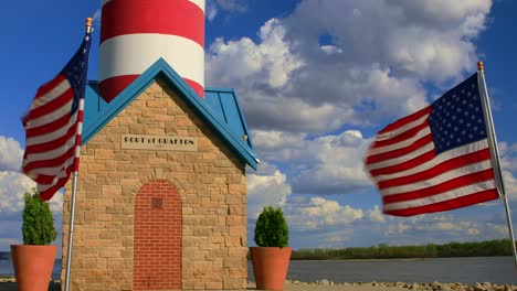 Faro-Del-Puerto-De-Grafton-Con-Banderas-Americanas-Ondeando-En-El-Viento-Contra-El-Cielo-Azul-Y-Las-Nubes,-Illinois,-EE.UU.