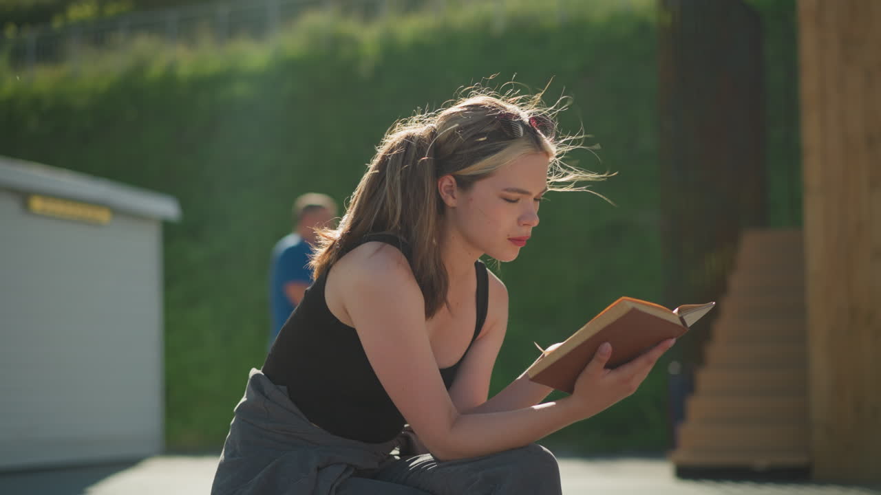 Lady seated outdoors reads a book while the wind blows through her hair and book pages, with sunlight reflecting on her, blurred background includes a staircase and a man passing by in a blue top