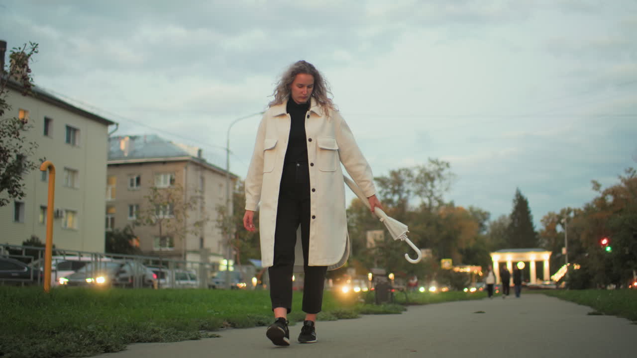 Woman in white coat walking with umbrella swinging in hand, residential buildings, blurred moving cars with headlights, benches, trees, and distant people under soft evening sky in calm urban setting