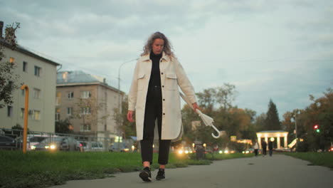 woman in white coat walking with umbrella swinging in hand, residential buildings, blurred moving cars with headlights, benches, trees, and distant people under soft evening sky in calm urban setting