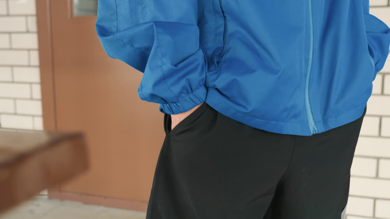 blue jacket closeup hand on pocket athlete standing in tiled corridor beside wooden bench and brick wall, casual windbreaker zipper visible, hand gestures and pocket interaction across five short