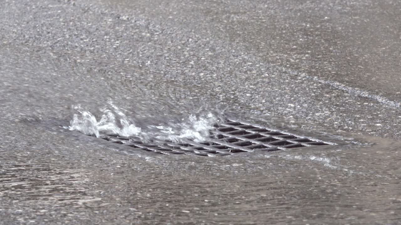 Water flows into a street drain as cars drive by.