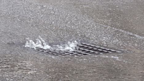 water flows into a street drain as cars drive by.