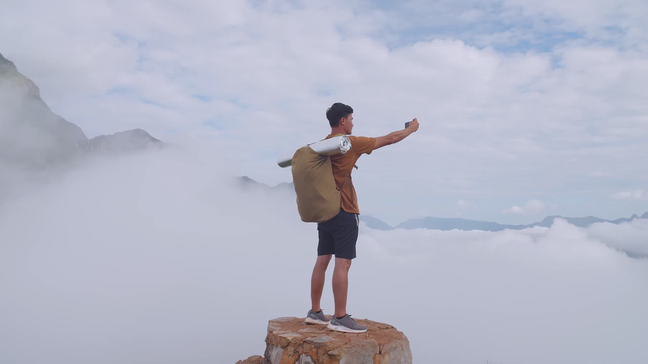 스마트워치를 이용해 산 위에 서서 안개가 가득한 산의 사진을 찍는 아시아인 하이커