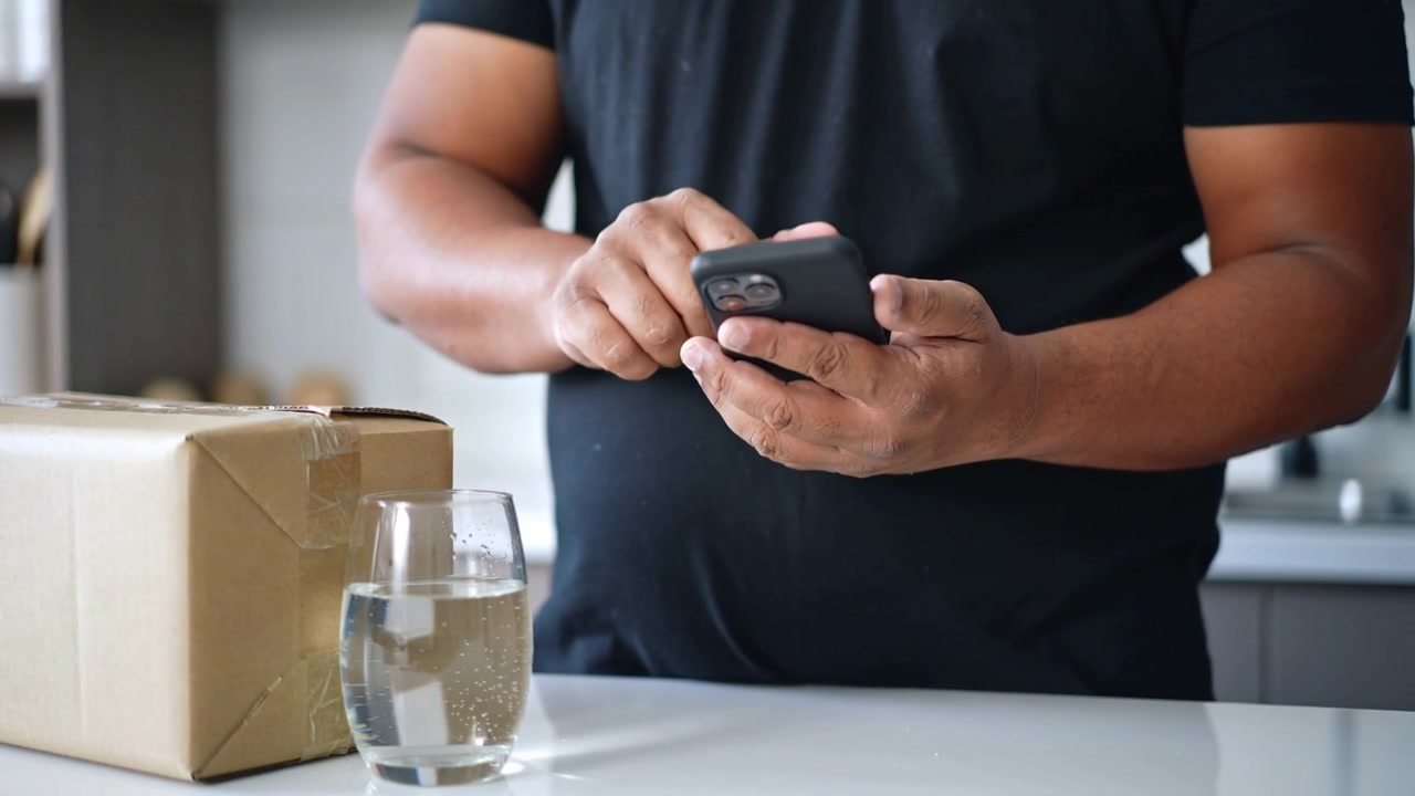 Checking phone for package status, man in black tee tapping and swiping at kitchen counter, glass