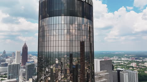 aerial descending shot of mirror facade of modern cylindrical skyscraper reflecting surrounding buildings and clouds in sky