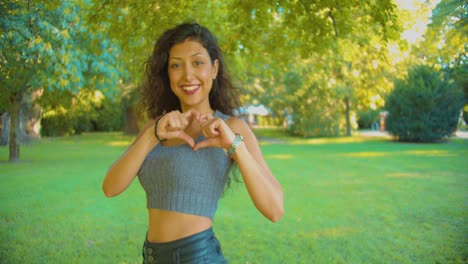 Joven-Atractiva-Mujer-Del-Medio-Oriente-Besando-La-Cámara-Y-Mostrando-El-Signo-De-La-Mano-Del-Corazón-En-El-Exterior-Del-Parque-Público,-Cámara-Lenta